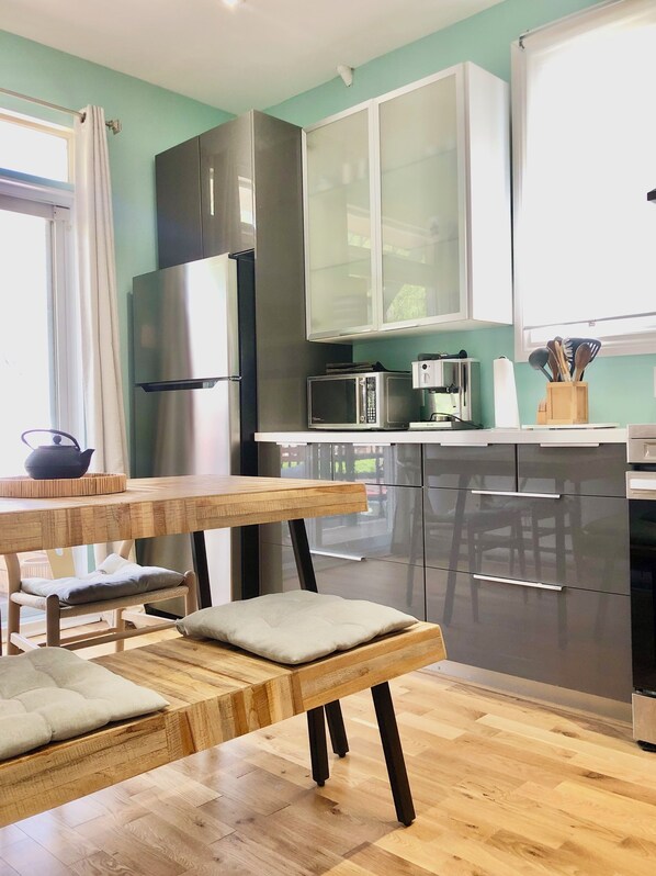 Cocina moderna con un refrigerador de acero inoxidable, un microondas y una cafetera. Hay una mesa de madera con un banco y cojines.
