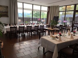 A dining area with multiple tables set for service, large windows offering a view of greenery, and a modern interior design.