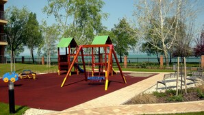 Un parque infantil con columpios y toboganes, rodeado de árboles y vistas al mar.