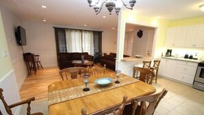 A dining area with a wooden table, chairs, and a chandelier. There is a sofa set and a kitchen area with cabinets and appliances.