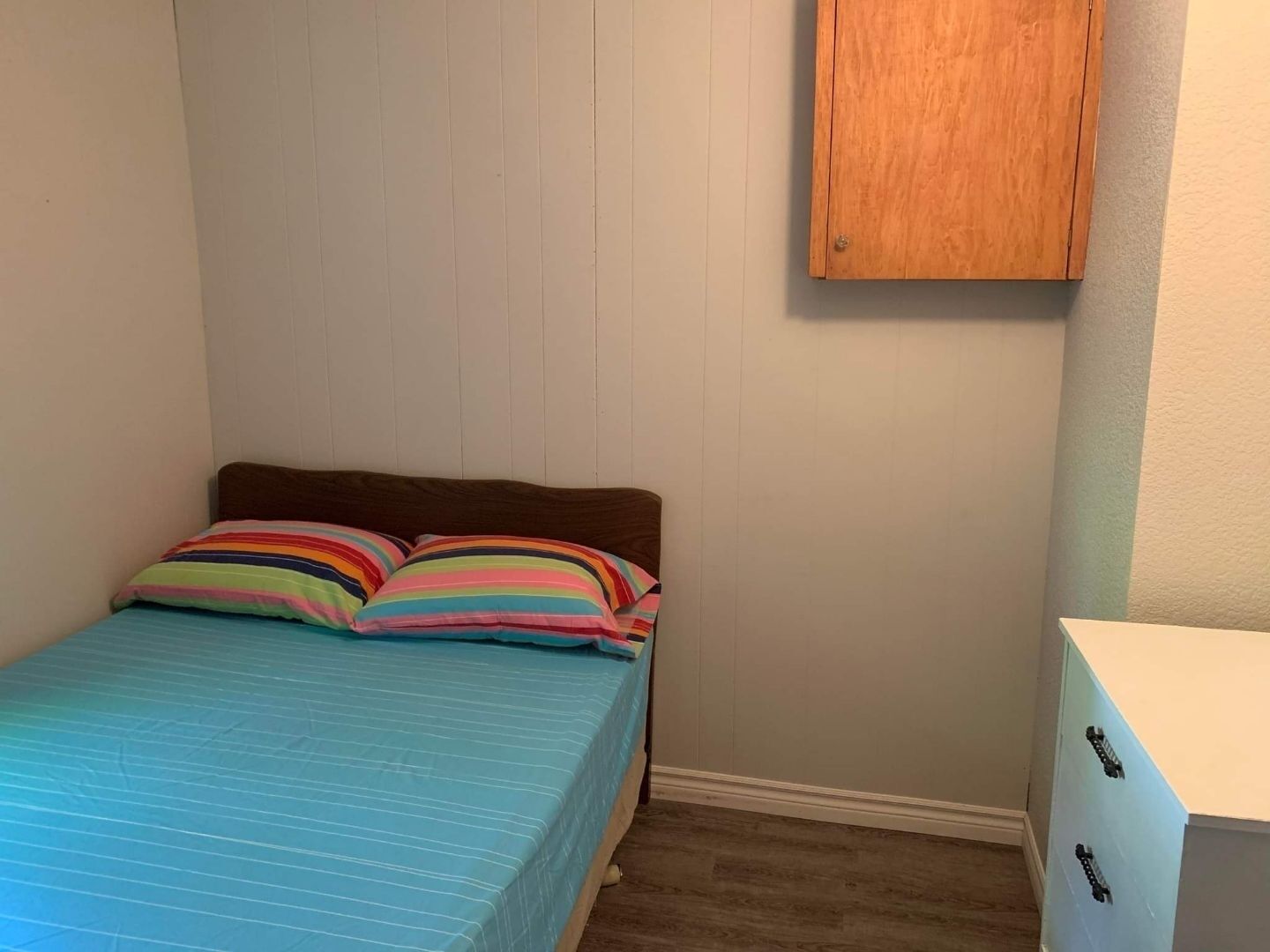 Une petite chambre à coucher, bien rangée, avec un lit simple, des coussins colorés à rayures, une tête de lit en bois et une commode blanche avec des tiroirs.