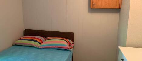 Une petite chambre à coucher, bien rangée, avec un lit simple, des coussins colorés à rayures, une tête de lit en bois et une commode blanche avec des tiroirs.