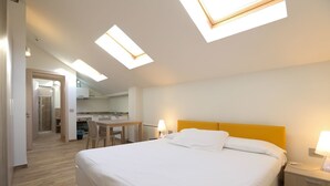 A hotel room with a bed, bedside table, and a small desk with a chair. There is a skylight above the bed and a separate skylight above the desk. - Amalia Bakery Home (Gallarate)