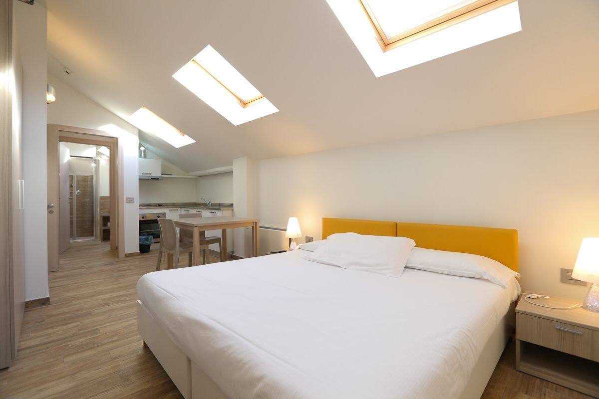 A hotel room with a bed, bedside table, and a small desk with a chair. There is a skylight above the bed and a separate skylight above the desk.
