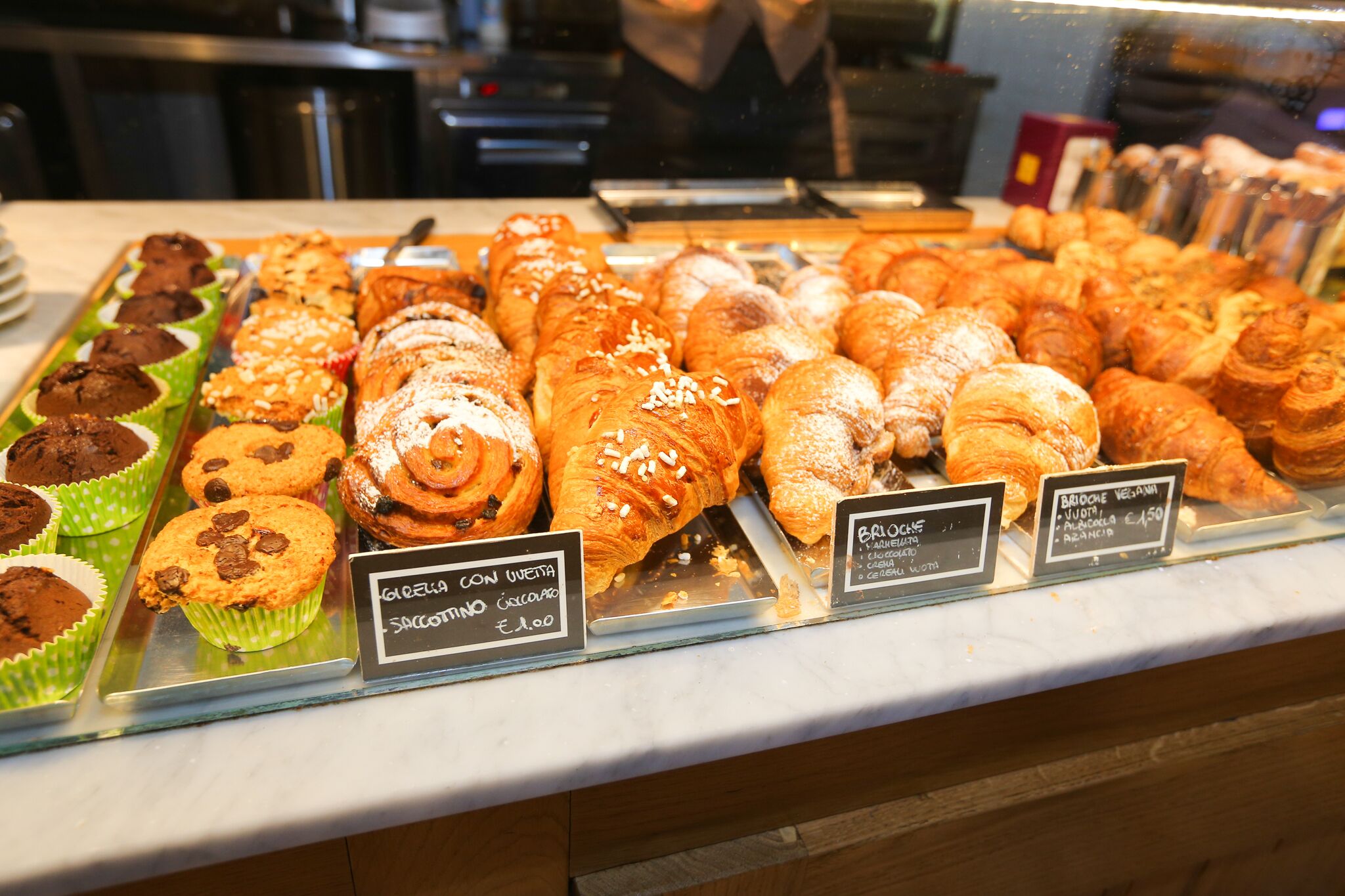 Mostra de forn amb diverses pastes i muffins, incloent muffins de xocolata i croissants, amb preus indicats.