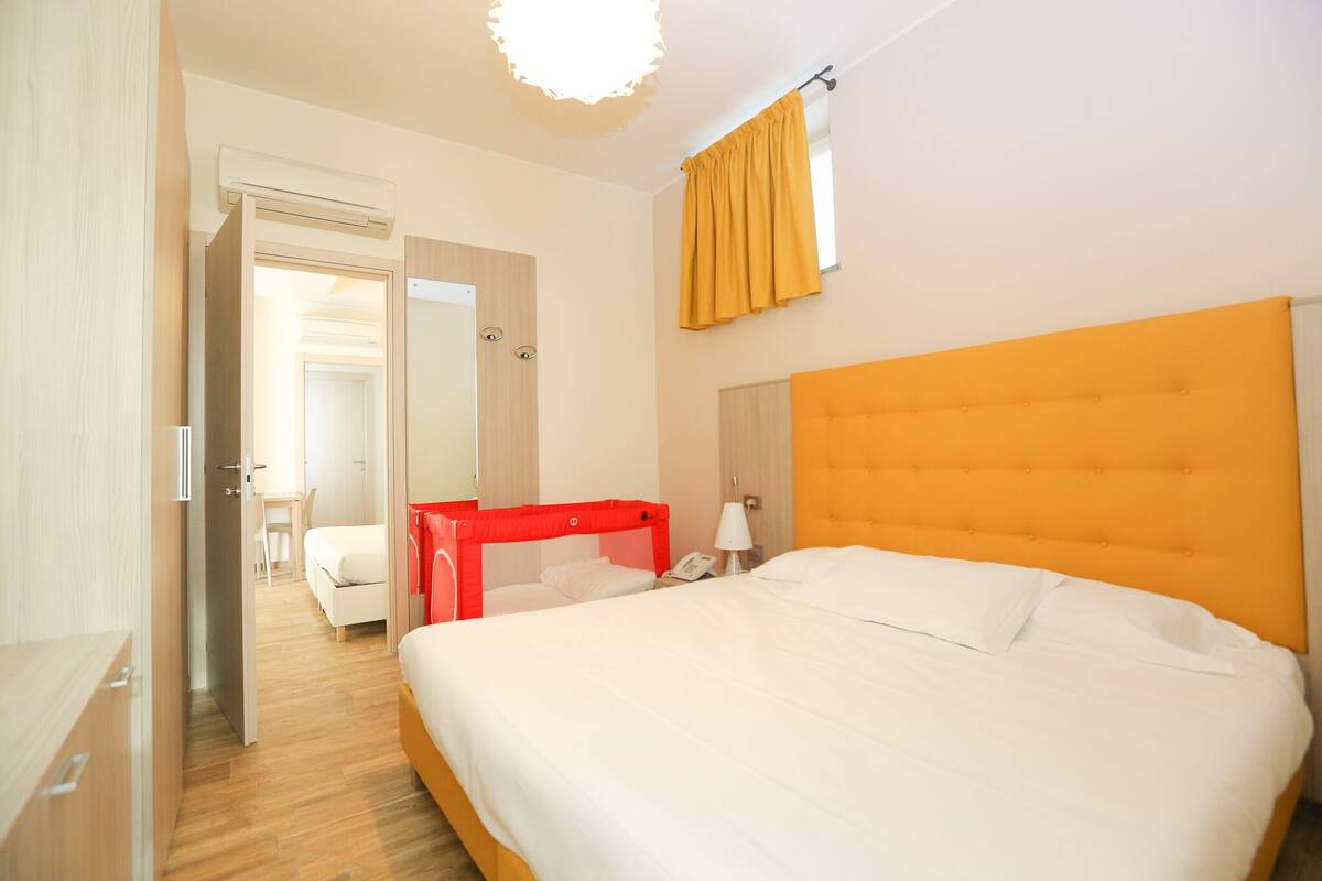 A hotel room with a large bed, a red headboard, a bedside table with a lamp, and a window with yellow curtains.