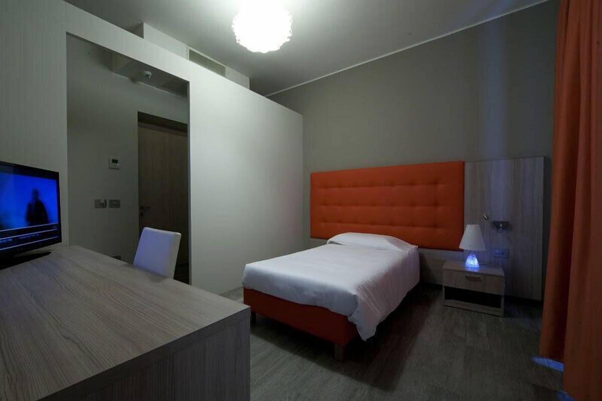 A hotel room with a bed, a desk with a television, a nightstand with a lamp, and a door leading to another room.