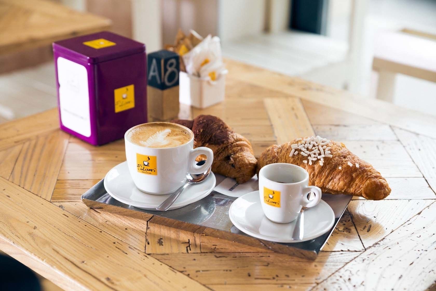 Et træbord med to hvide kaffekopper, en croissant med hvide drys og en lilla æske med teksten "A18".