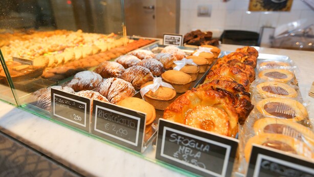 A bakery display case with various pastries and desserts, including a long loaf of bread, macarons, and other baked goods. - Amalia Bakery Home (Gallarate)