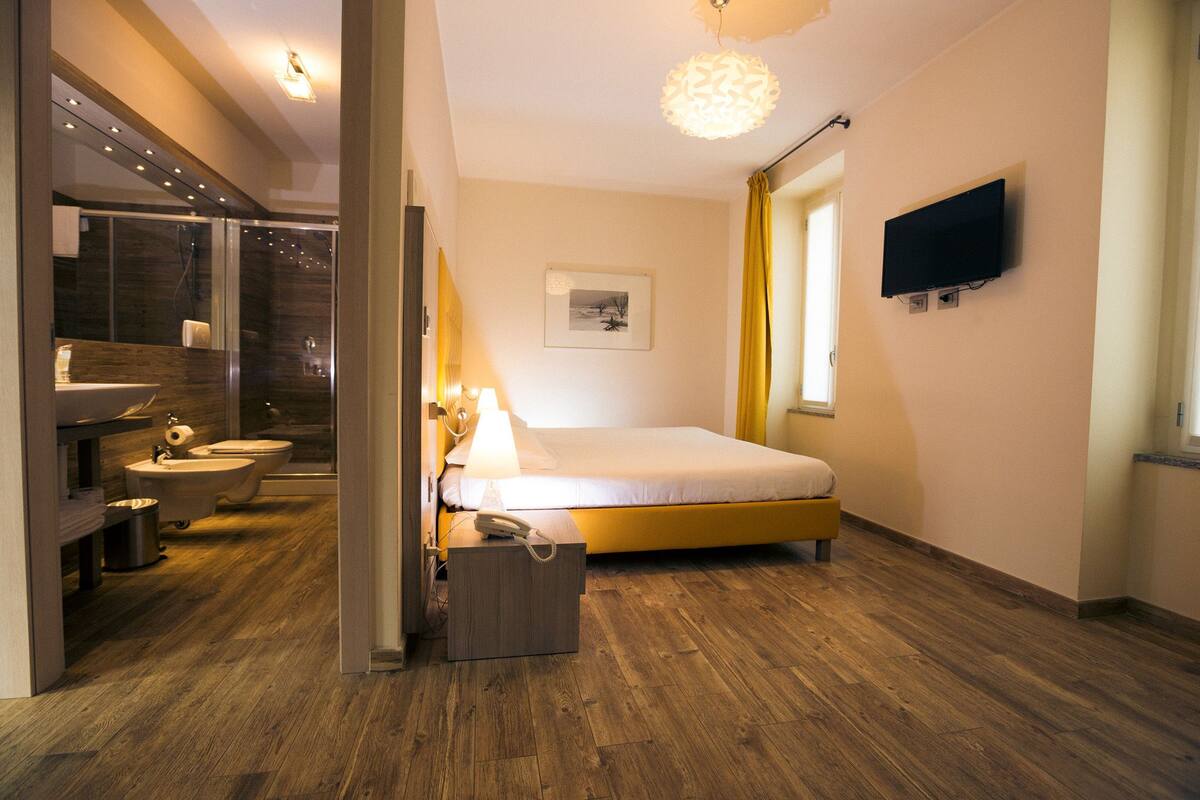 A modern hotel room with a wooden floor, a bed, a bedside table with a lamp, a TV mounted on the wall, and a bathroom with a shower, sink, and toilet.