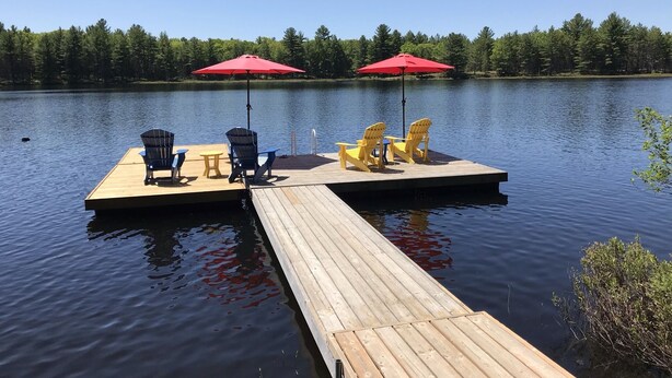 Une jetée en bois avec des chaises et des tables, un parasol rouge et un lac paisible, avec des arbres en arrière-plan.