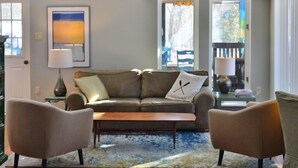 Un salon avec un plafond haut, une grande fenêtre, un canapé, deux fauteuils, une table basse et un tapis.