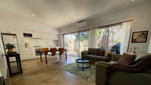 A modern living room with a sofa, a coffee table, and a dining area with chairs and a table. There is a kitchen in the background with white cabinets and a microwave.