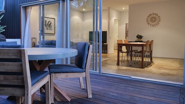 A modern interior with a dining area featuring a round table and chairs, a wooden deck, and a sliding glass door leading to a living space.