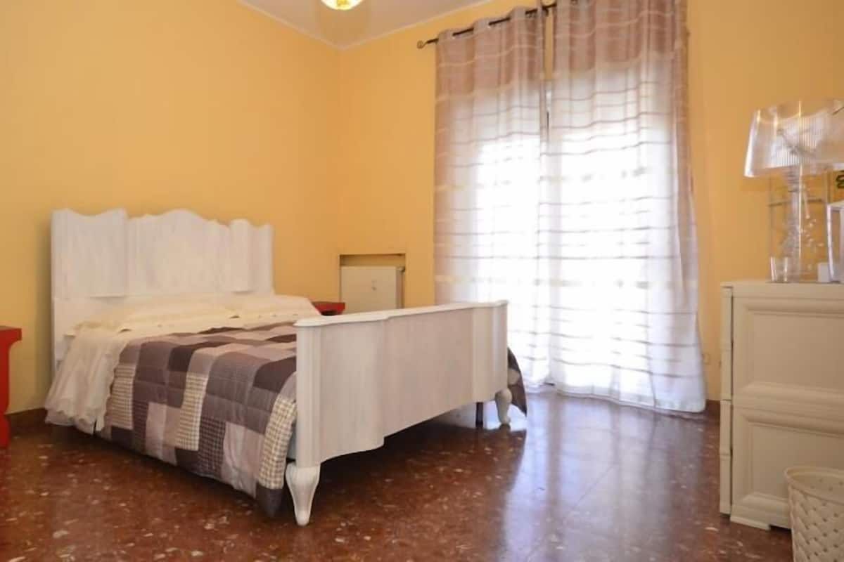 Un dormitorio con una cama, un taburete pequeño rojo, una mesita de noche y un ventanal con cortinas.