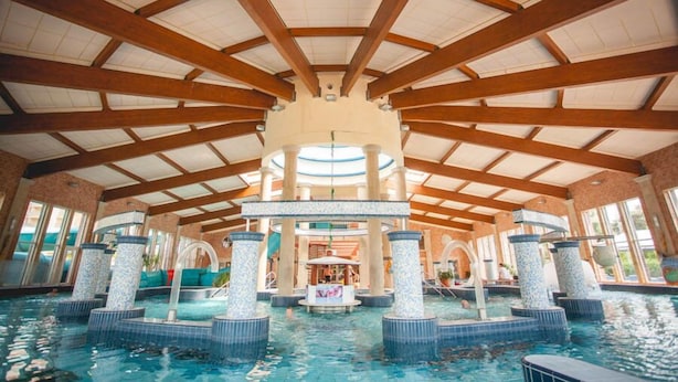 An indoor swimming pool with a high ceiling and wooden beams, featuring multiple water fountains and a central play area. - Galerius Panorama Beach - D Apartman (Siófok)