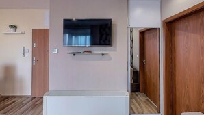 A modern living room with a flat-screen TV mounted on the wall, a built-in air conditioning unit below it, and a wooden door to the right. - Galerius Panorama Beach - D Apartman (Siófok)
