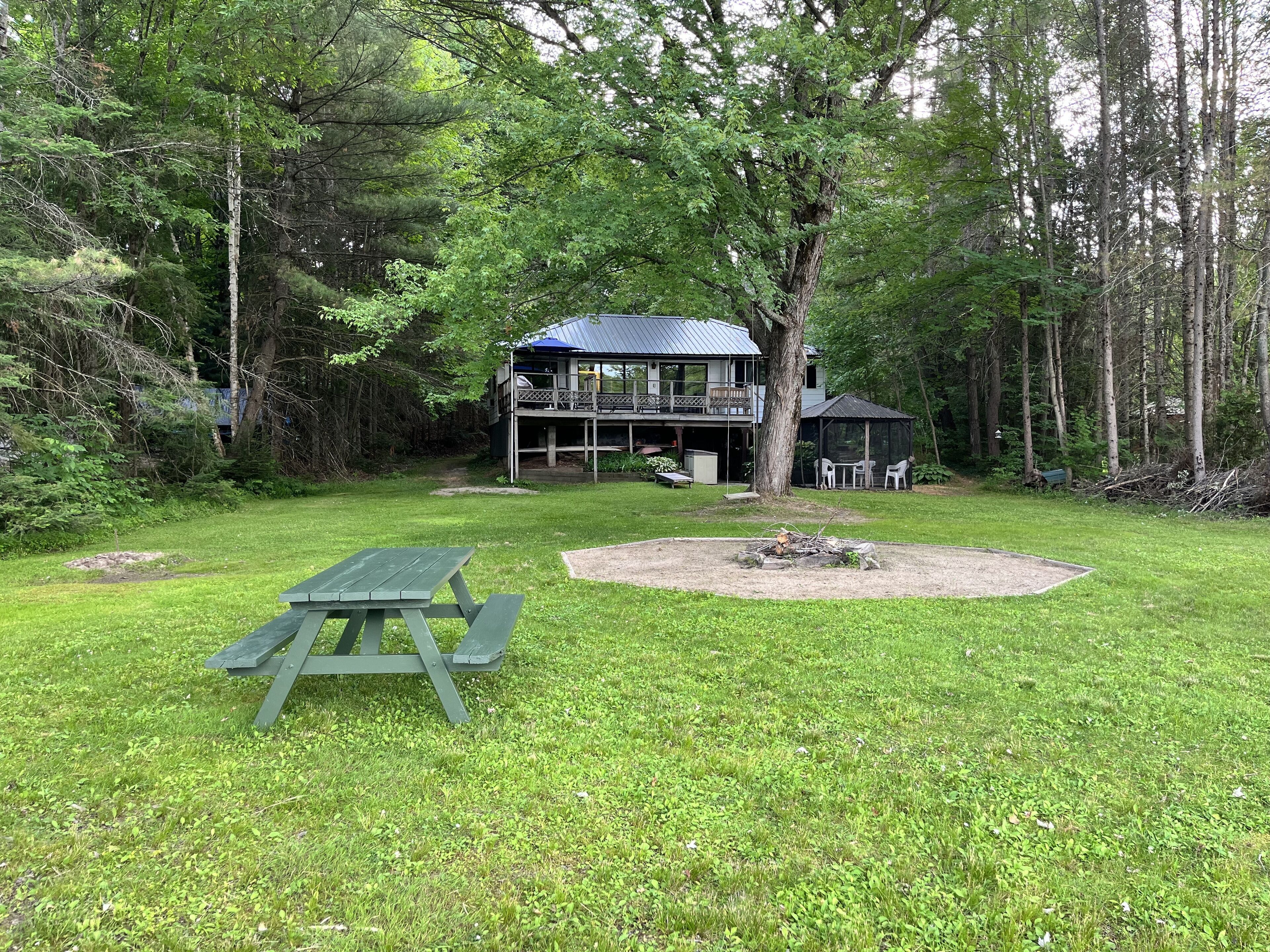 A cabin with a deck and a covered seating area, surrounded by a grassy yard and trees.