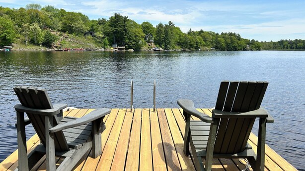 Un muelle de madera con dos sillas Adirondack mirando un lago tranquilo, rodeado de exuberante vegetación y edificios a la distancia.