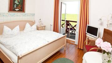 A bedroom with a bed, a TV, a chair, a table with a vase, and a view of a grassy area through an open door.