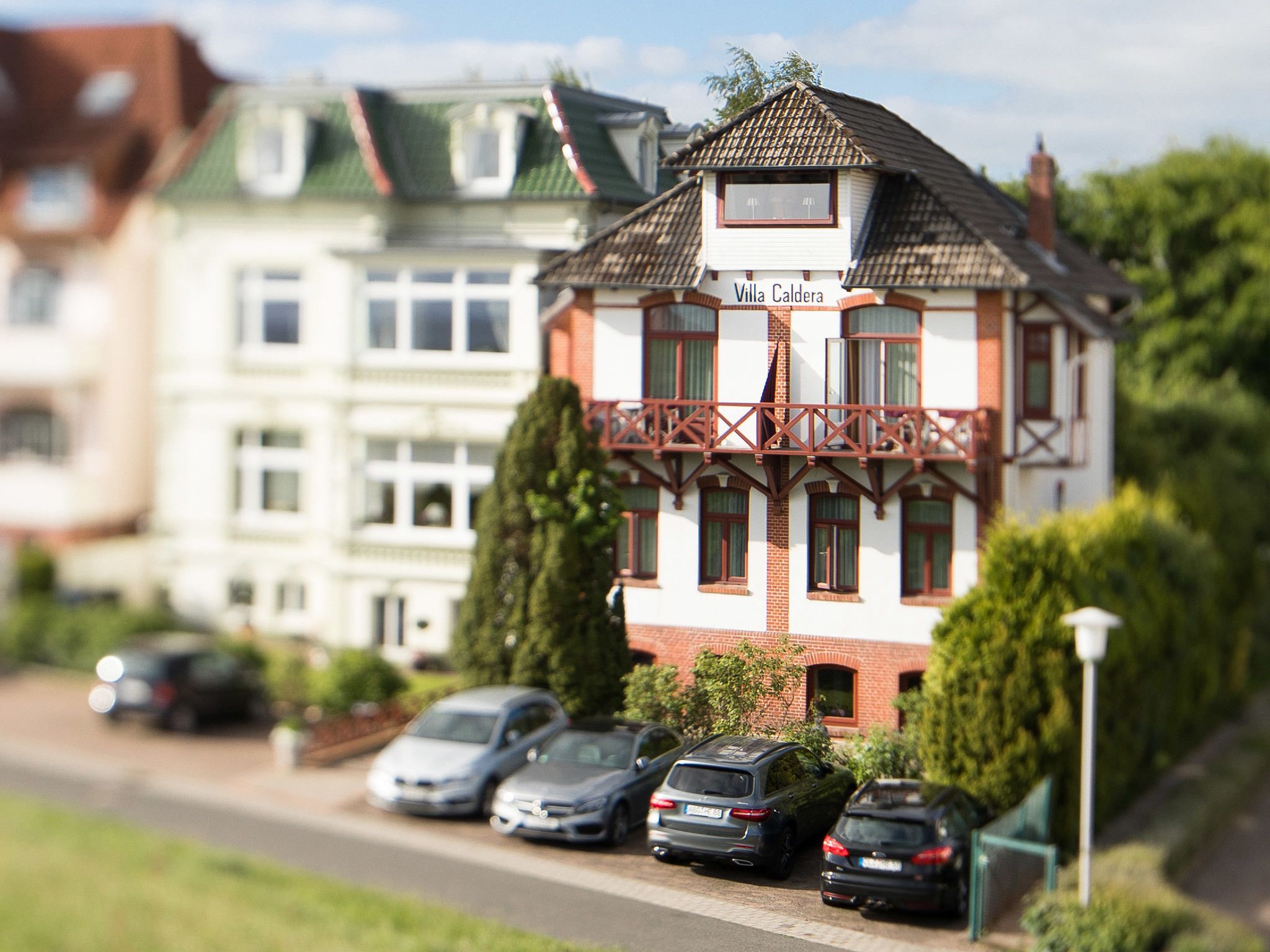 A model of Villa Caldera with a traditional European architectural style, featuring a balcony and a row of parked cars in front.