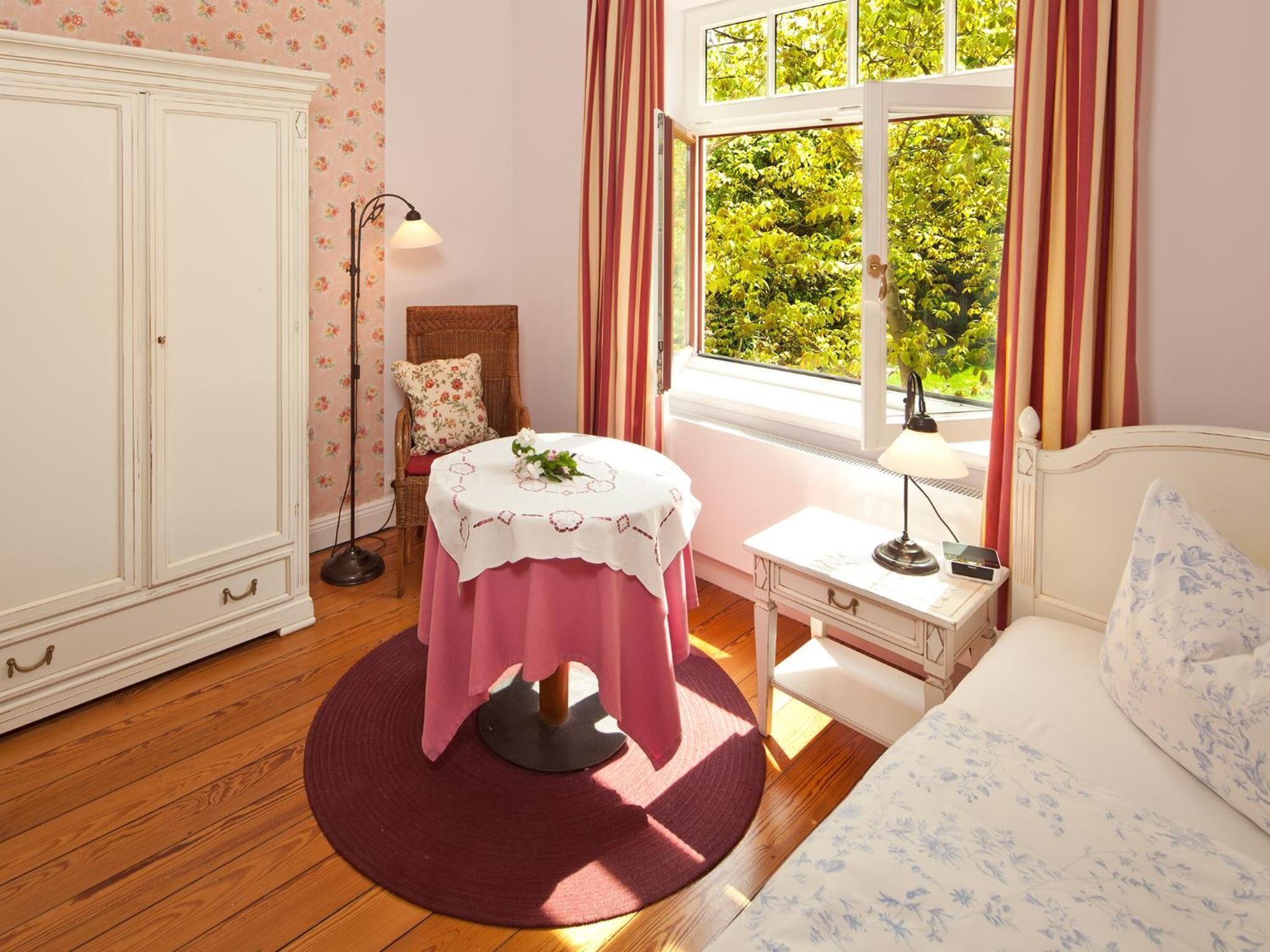 Une pièce comprenant un lit, un bureau avec une lampe, une chaise en osier, une table recouverte d’une nappe et une armoire.