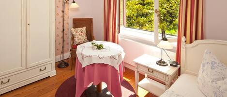 Une pièce comprenant un lit, un bureau avec une lampe, une chaise en osier, une table recouverte d’une nappe et une armoire.