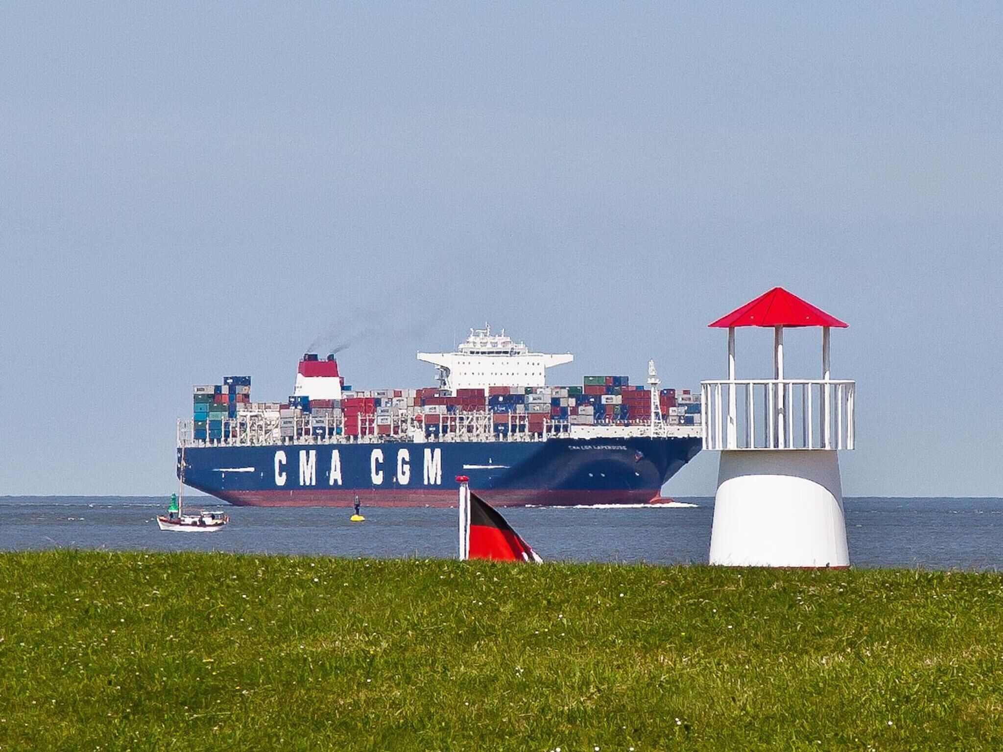 A large cargo ship with the text "CMA CGM" on its side, docked near a grassy area with a small boat and a red and white structure.