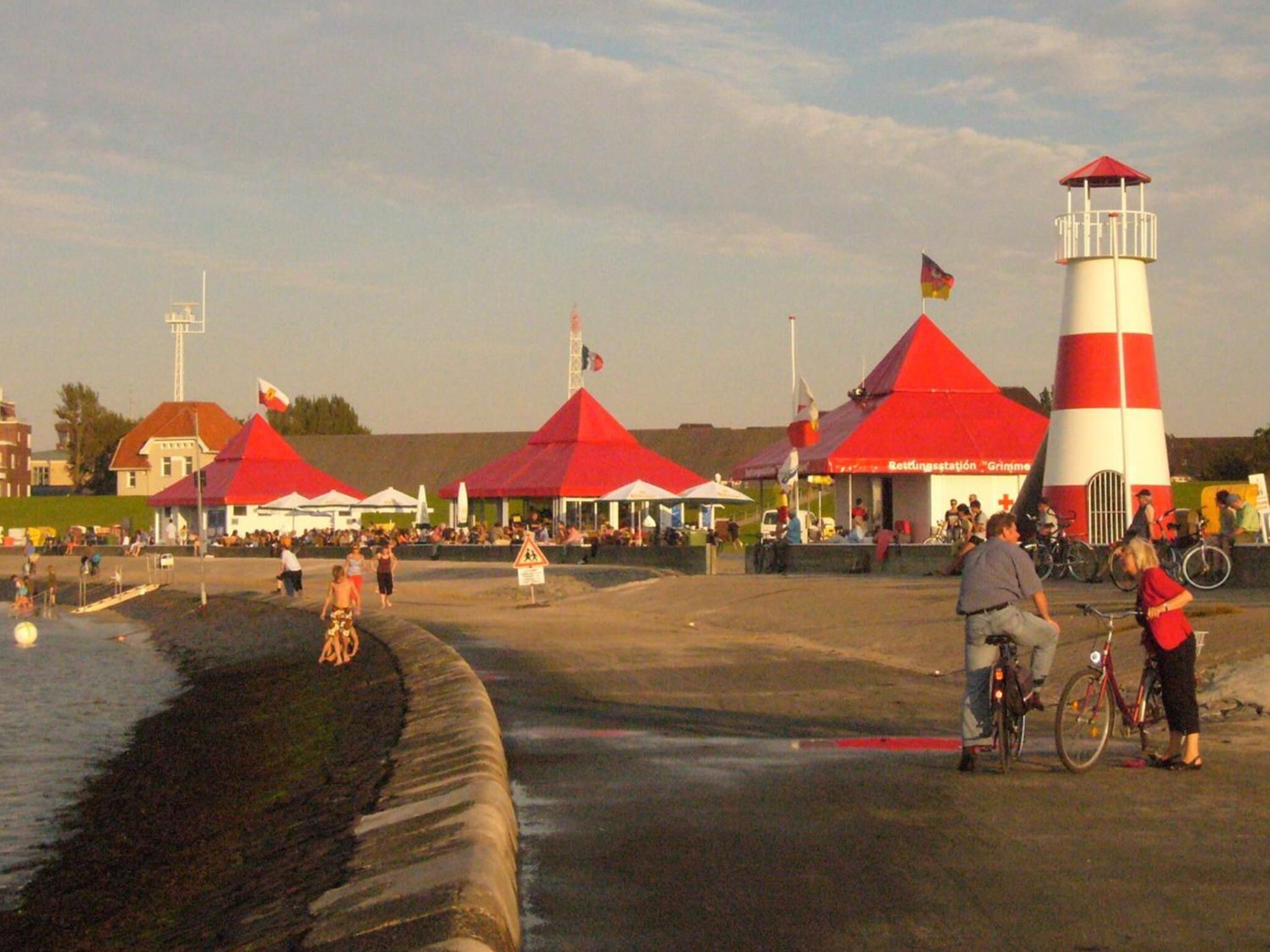 Zona costera con un faro, tiendas de campaña rojas y una playa de arena.