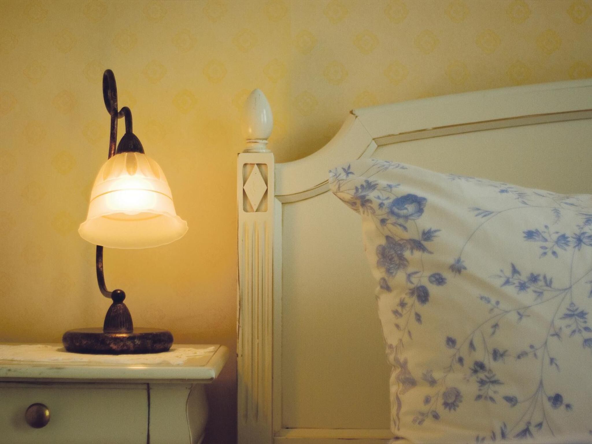 A bedside table with a lamp, a bed with a floral bedspread, and a patterned wall.