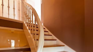 Una escalera con balaustres blancos y peldaños de madera, que conduce a un rellano con una lámpara fijada a la pared.