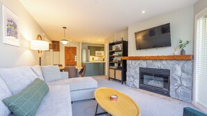 A living room with a sofa, a coffee table, a fireplace, a TV, and a kitchen in the background. - Aspens #358 Prime Ski-in Ski-out Location! Pool, Hot tubs, BBQ, sleeps 6 (Whistler)