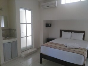 Habitación de hotel con cama, mesita de noche, pequeño tocador, baño con lavamanos y espejo, y una ventana.