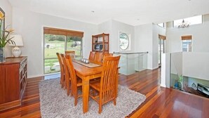 Un comedor amplio con una mesa y sillas de madera, un tapete blanco y ventanas grandes que ofrecen vistas al exterior.
