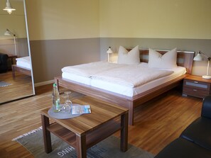 Une chambre d’hôtel bien rangée, avec un lit en bois, des tables de chevet et une petite table sur laquelle se trouvent une bouteille et des verres.