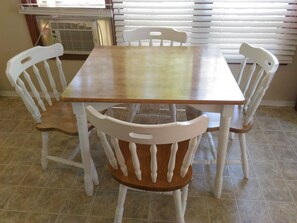 Une table à manger avec quatre chaises blanches, une fenêtre avec des stores et un appareil de climatisation.