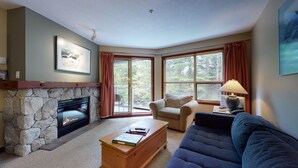 A cozy living room with a stone fireplace, a large window with curtains, a blue sofa, and a wooden coffee table. - Aspens #216 Prime Ski-in Ski-out Location! Pool, Hot tubs, BBQ, sleeps 4 (Whistler)