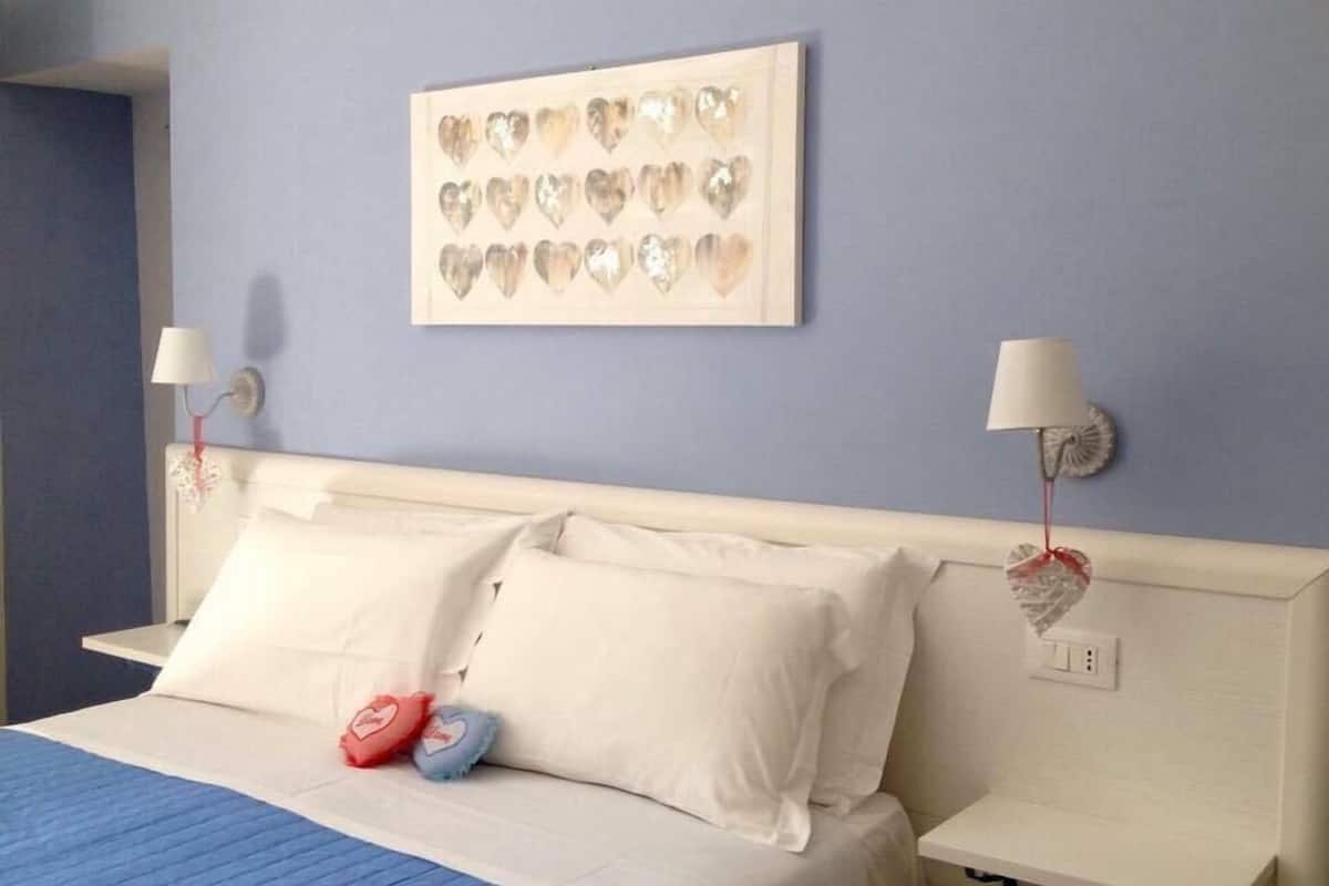 A neatly made bed with white linens and blue bedding, a wall-mounted heart-shaped light fixture, and a framed picture of hearts on the wall.