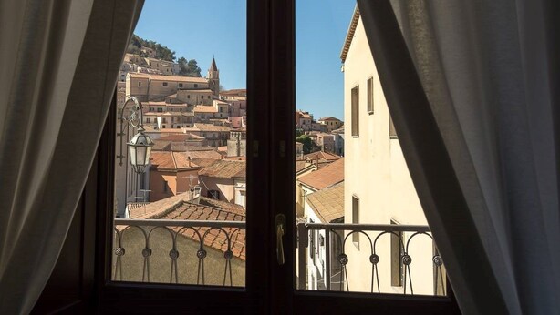 Une vue depuis une fenêtre, à travers des rideaux, donnant sur une zone urbaine densément peuplée, avec des bâtiments historiques et le clocher imposant d’une église.