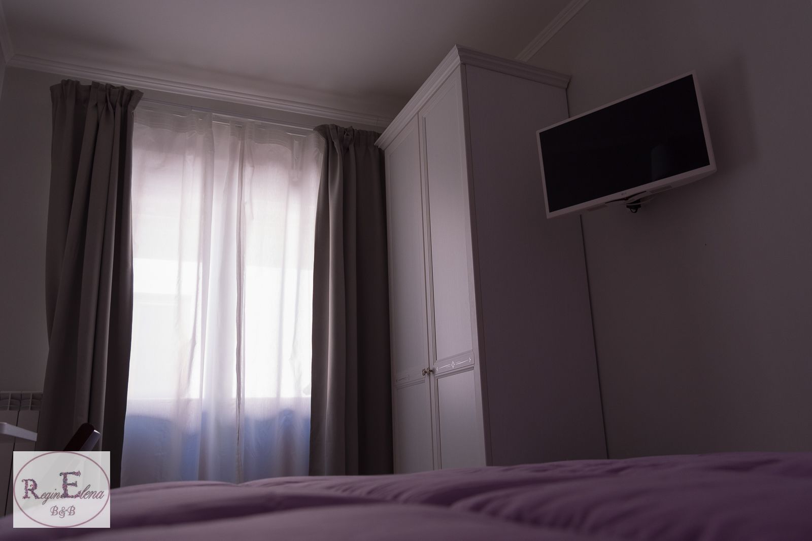 Un dormitorio con cama, armario, televisor montado en la pared y ventana con cortinas.