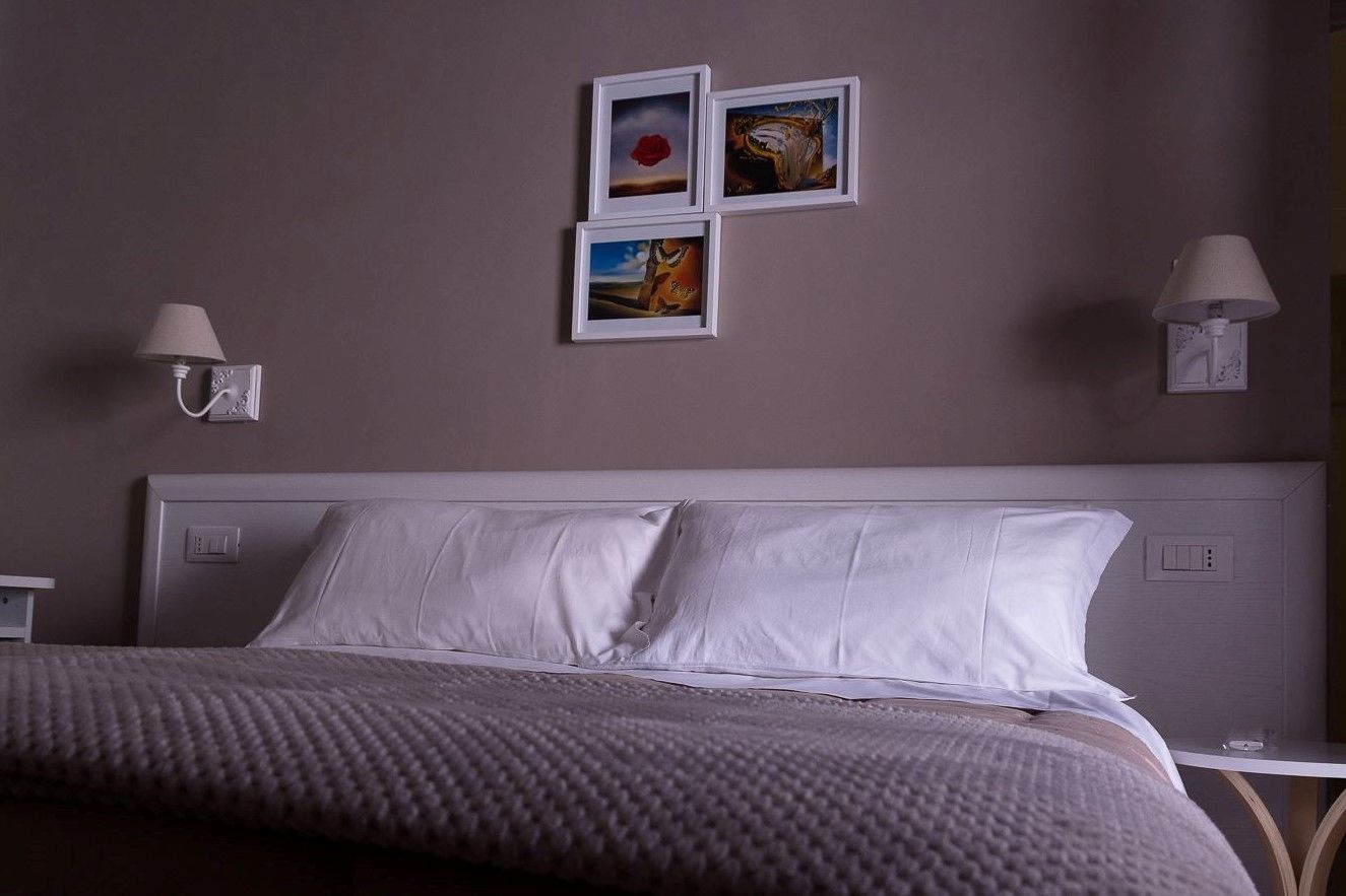 Una cama bien hecha con sábanas blancas, un plaid gris texturizado y un cabecero con un interruptor de luz. Hay tres cuadros enmarcados en la pared sobre la cama, y dos lámparas de pared a cada lado.