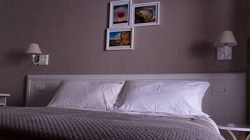 Una cama bien hecha con sábanas blancas, un plaid gris texturizado y un cabecero con un interruptor de luz. Hay tres cuadros enmarcados en la pared sobre la cama, y dos lámparas de pared a cada lado.