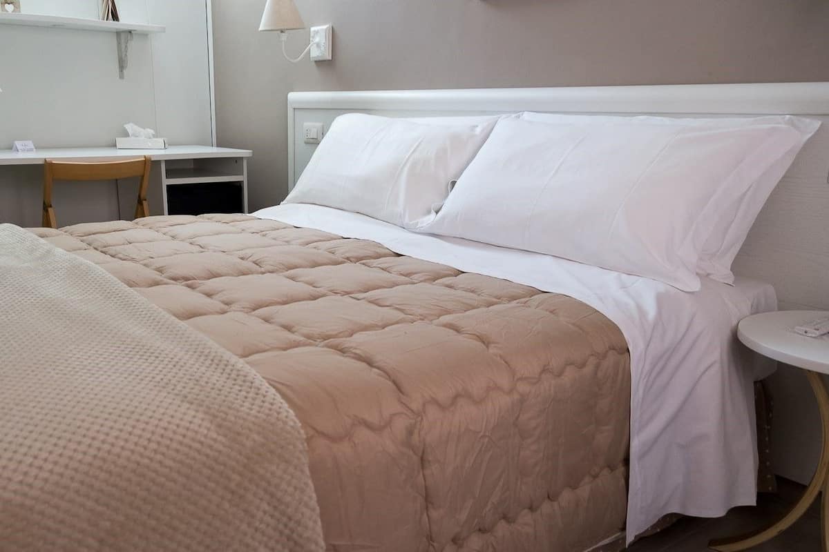 A neatly made bed with a beige quilt, a white pillow, and a white duvet. There is a small round table with a lamp beside the bed.
