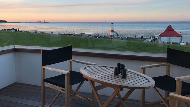 Un balcon avec une table et des chaises donnant sur une zone côtière, avec un phare et un bâtiment portant l’inscription « Rettungsstation ‚Grimmerhorn‘ ».