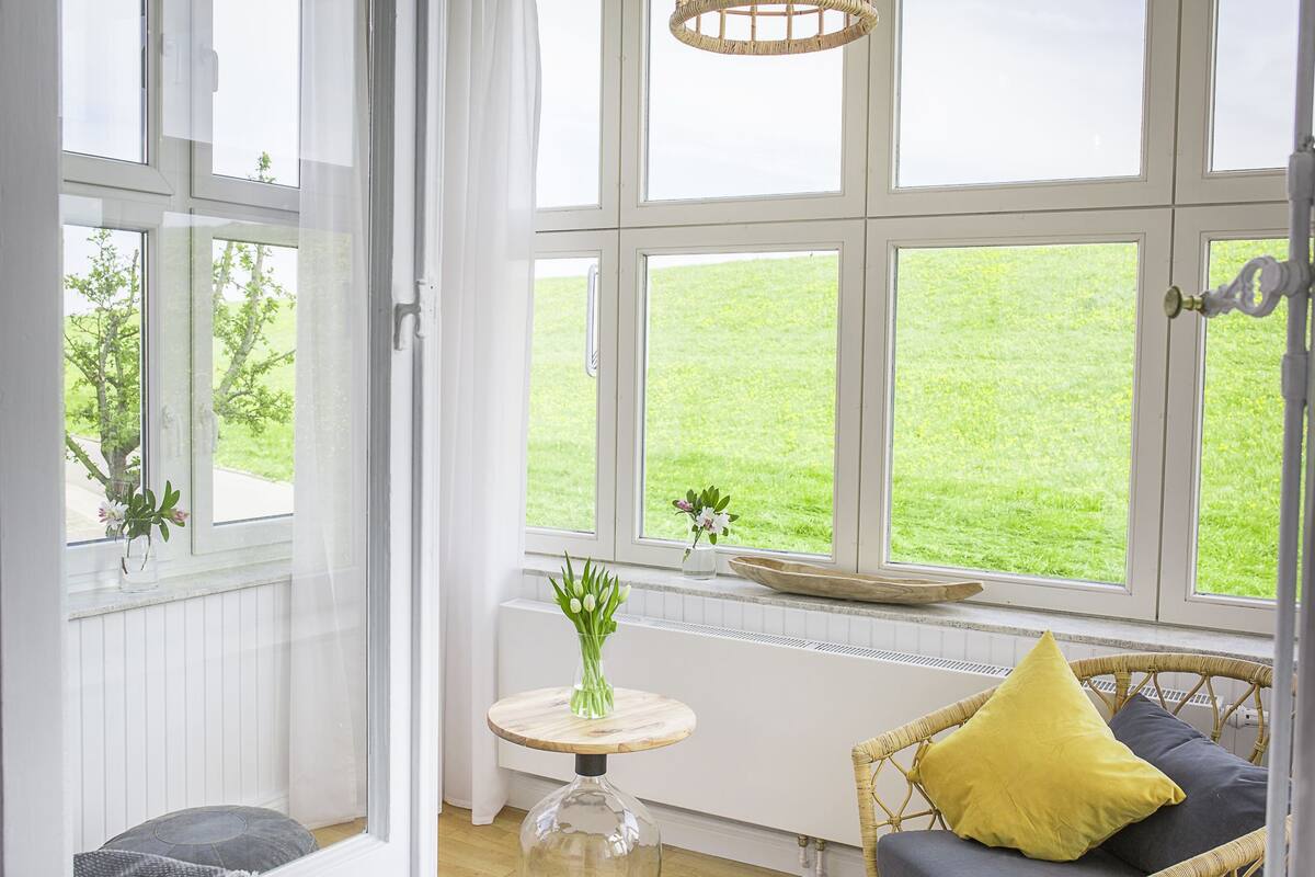 Et lyst rum med store vinduer, en fletstol, et lille bord med en vase og en radiator.
