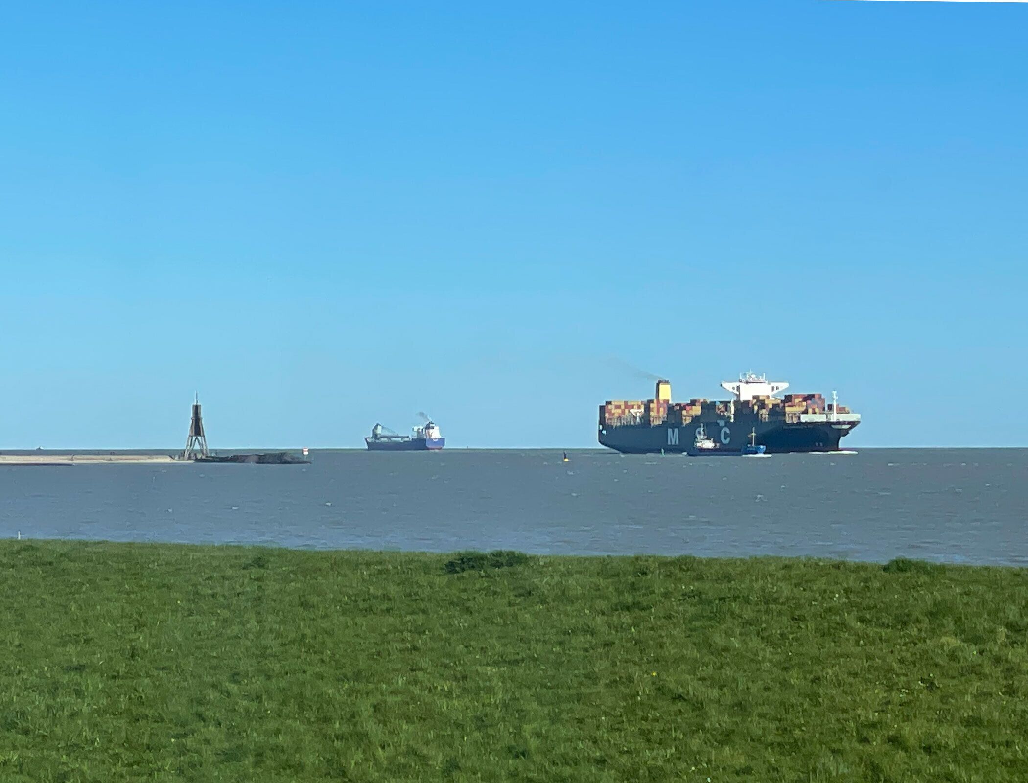 Um navio de carga com contêineres na água, um barco menor nas proximidades e uma margem gramada em primeiro plano.