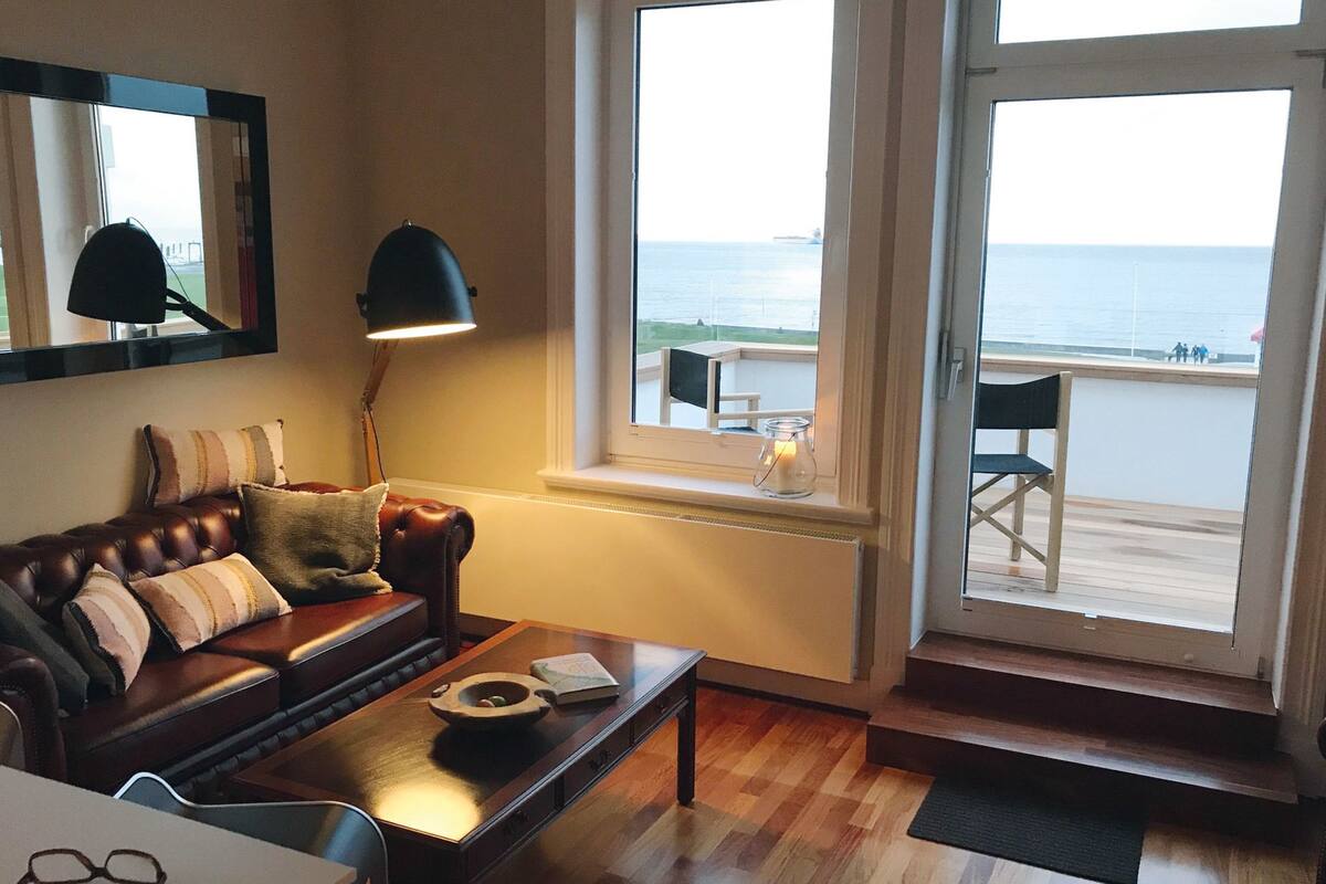 En hyggelig stue med en lædersofa, et træbord og havudsigt gennem store vinduer.