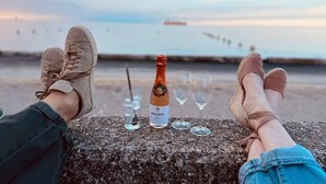 Une personne est assise sur une corniche en pierre, une bouteille de vin et des verres à portée de main, et elle a devant elle un plan d’eau avec une jetée visible au loin.