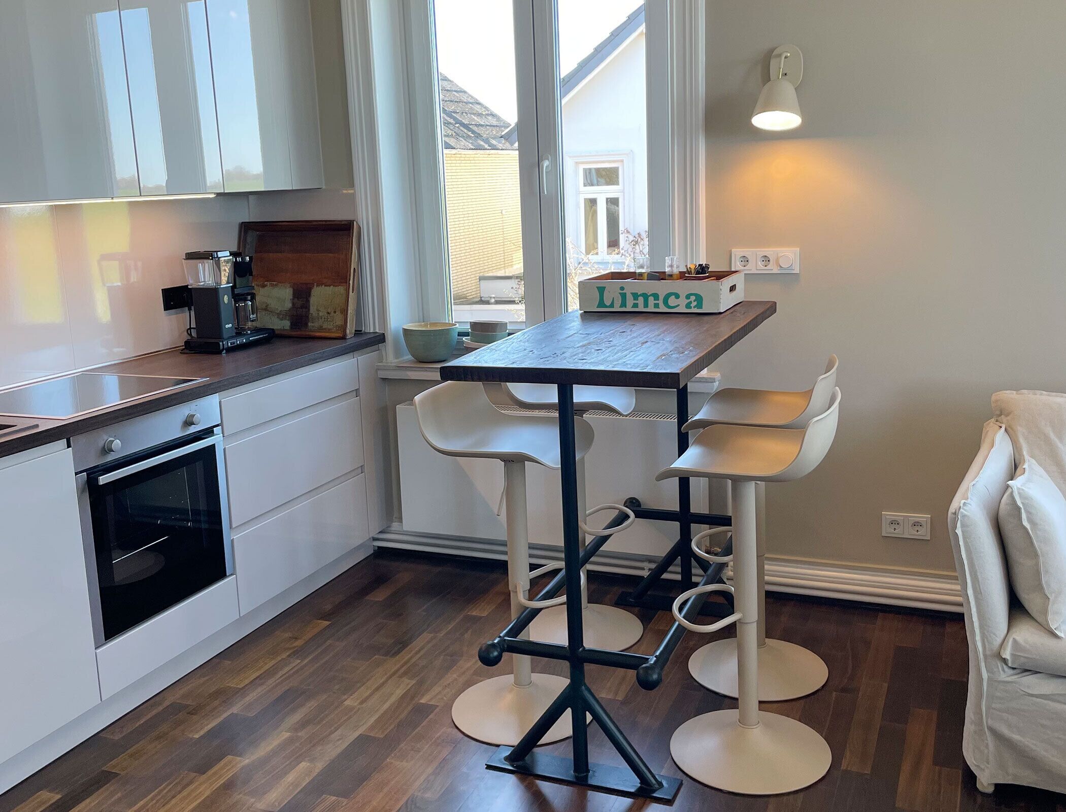 Une cuisine avec des meubles blancs, un four encastré, une machine à café et une table ornée d’une enseigne « Limca ».
