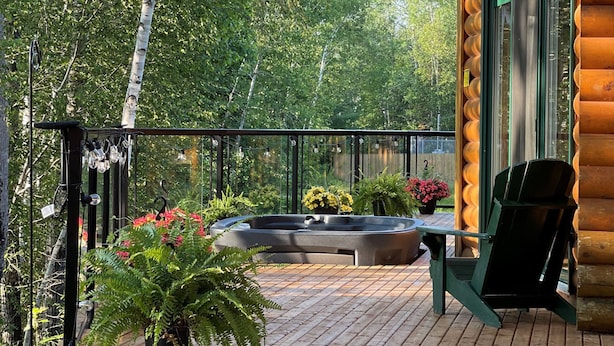 A wooden deck with a hot tub, surrounded by lush greenery and a black chair. - Red Bank Lodge - Red Bank, New Brunswick (Sillikers)